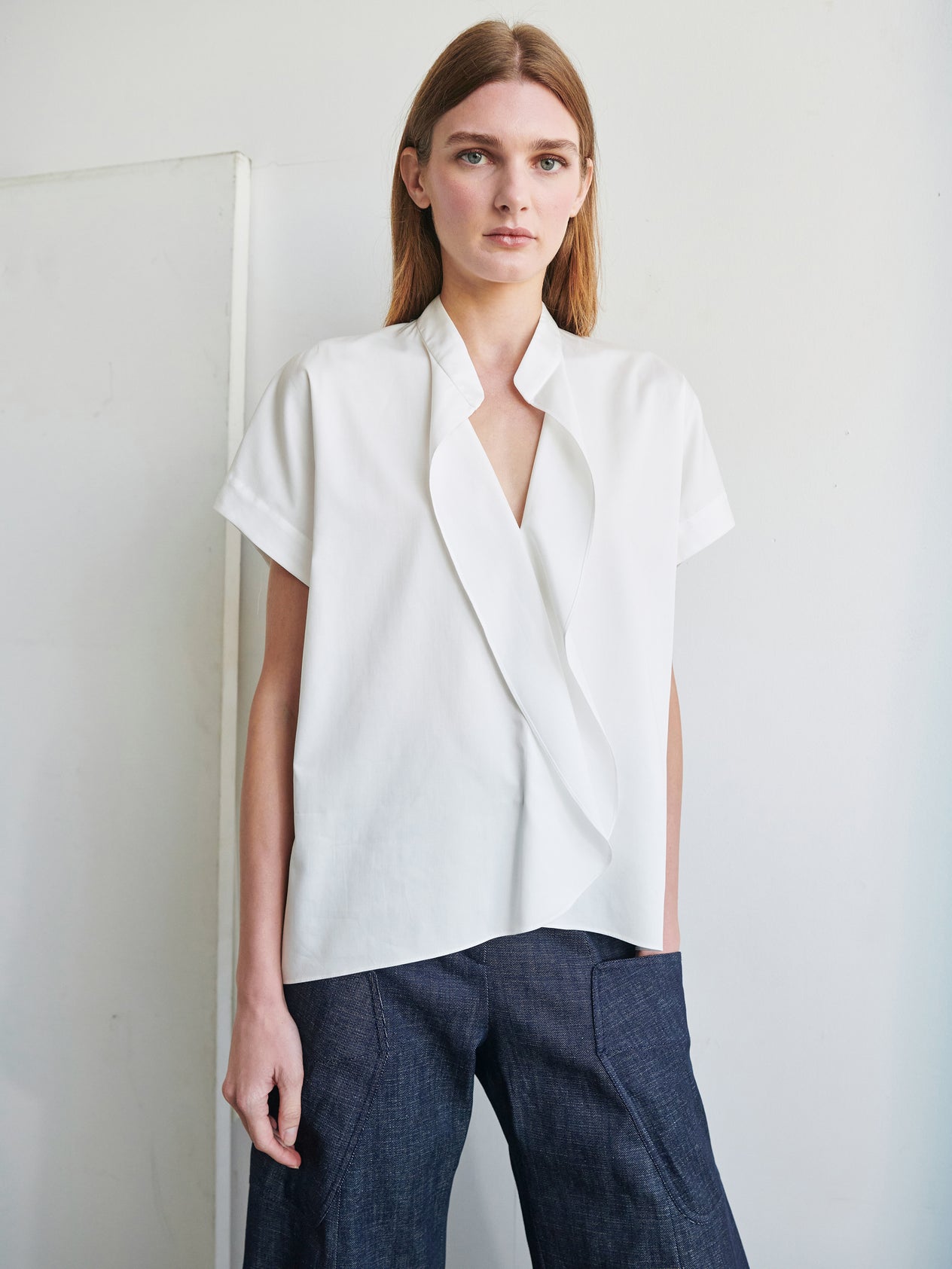 A woman with long brown hair stands against a plain white background. She is wearing a short-sleeved Fin Gaban Shirt by Zero + Maria Cornejo paired with light-colored pants, her hands casually in her pockets. She has a neutral expression and is looking directly at the camera.