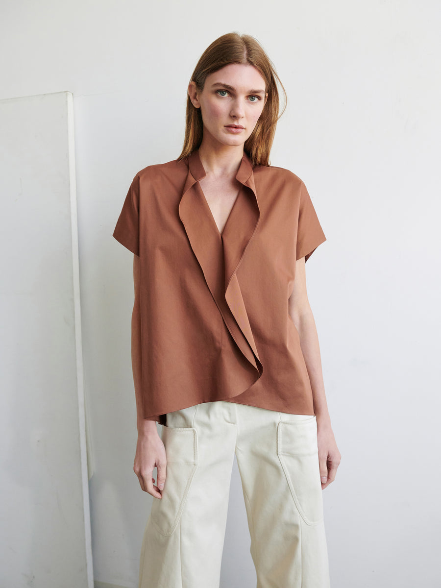 A woman with long, light brown hair stands facing the camera against a plain light-colored background. She is wearing Zero + Maria Cornejo's short-sleeved Fin Gaban Shirt with ruffle detail and light beige, high-waisted pants with front patch pockets. She has a neutral expression.