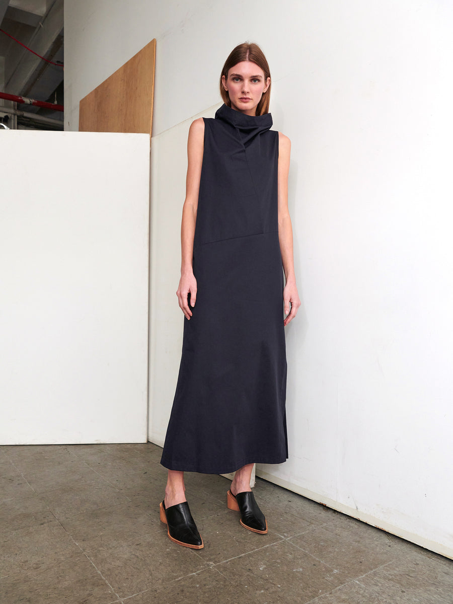 A person wearing the Long Fold Dress by Zero + Maria Cornejo, a sleeveless, dark blue cotton broadcloth piece, and black slip-on shoes stands against a white wall in an industrial-looking space. They have straight brown hair and maintain a neutral expression.