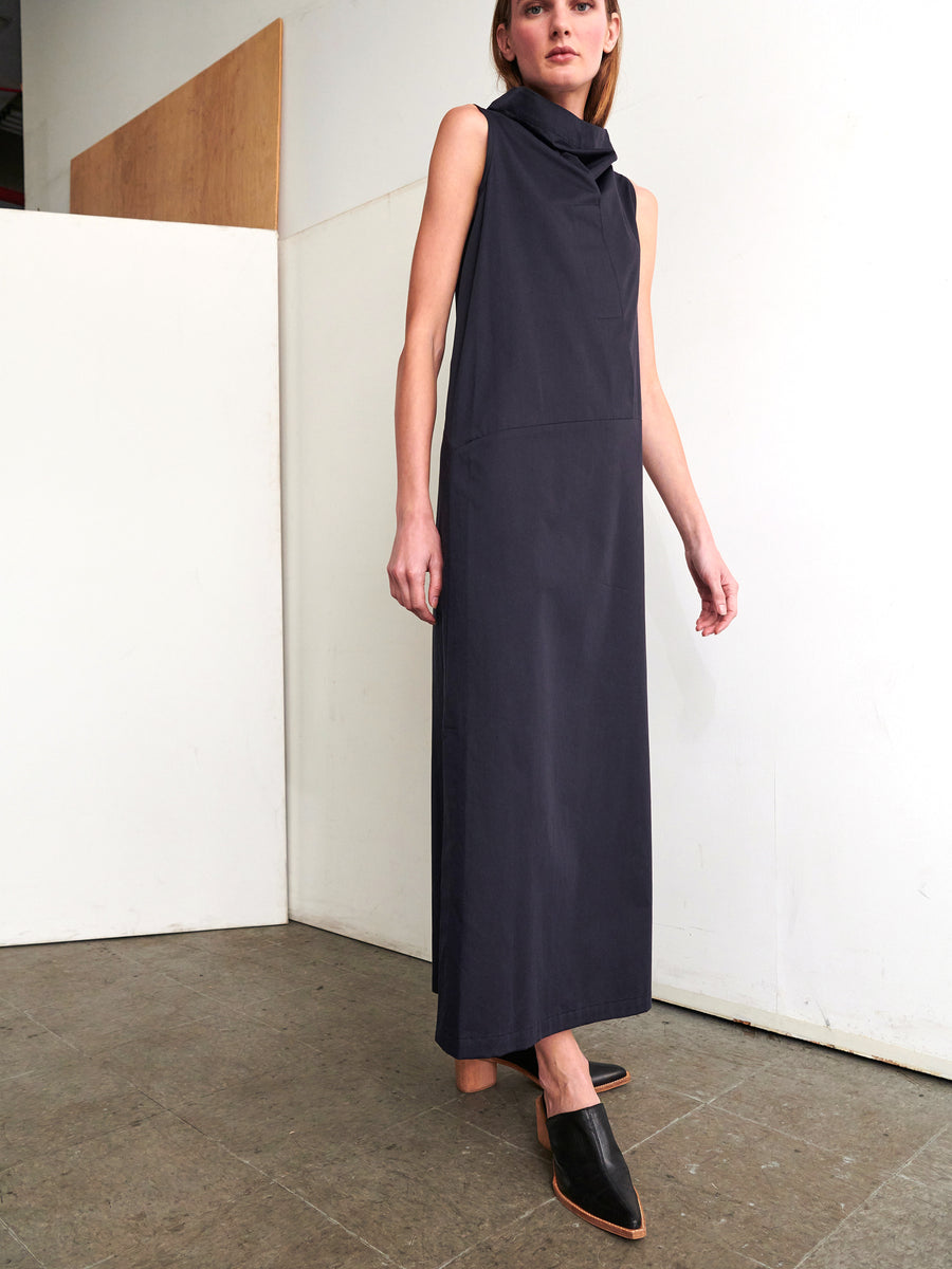 A person stands in a corner wearing the Long Fold Dress, a sleeveless, high-neck creation in cotton broadcloth by Zero + Maria Cornejo. The design is simple and elegant. They have short hair and are wearing a pair of black heeled mules. The setting is minimalistic, with neutral-colored walls and a tiled floor.