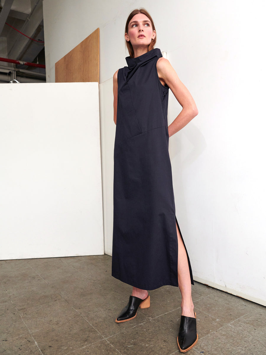 In a minimalist room, a person stands wearing the Long Fold Dress by Zero + Maria Cornejo. This sleeveless, mid-length dress features a high collar and side slit, crafted from dark cotton broadcloth. They have paired it with black mule shoes. Their long hair complements the plain wall backdrop that showcases a portion of exposed wood.