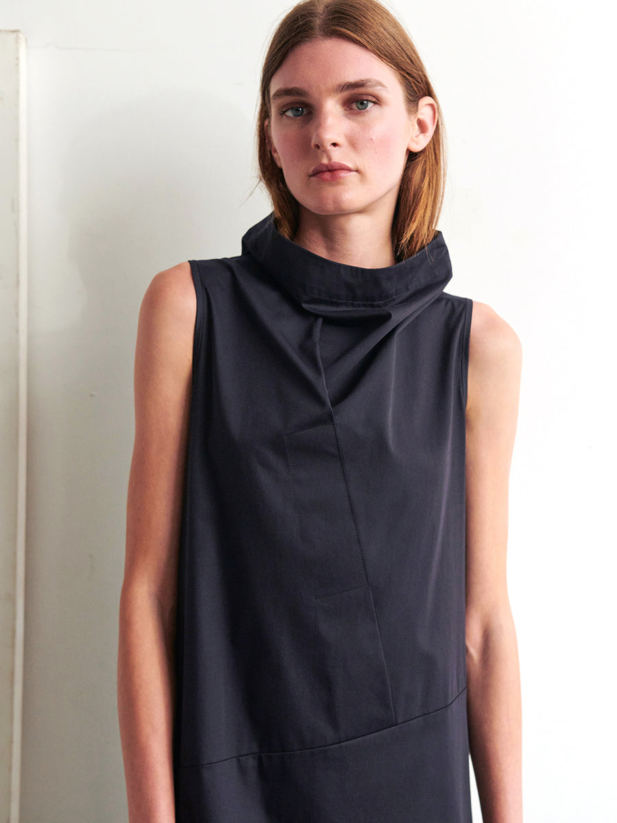 A woman with shoulder-length hair wears the Long Fold Dress by Zero + Maria Cornejo, a sleeveless, high-neck, dark cotton dress featuring an asymmetrical detail. She stands against a plain, light-colored wall, gazing directly at the camera with a neutral expression.