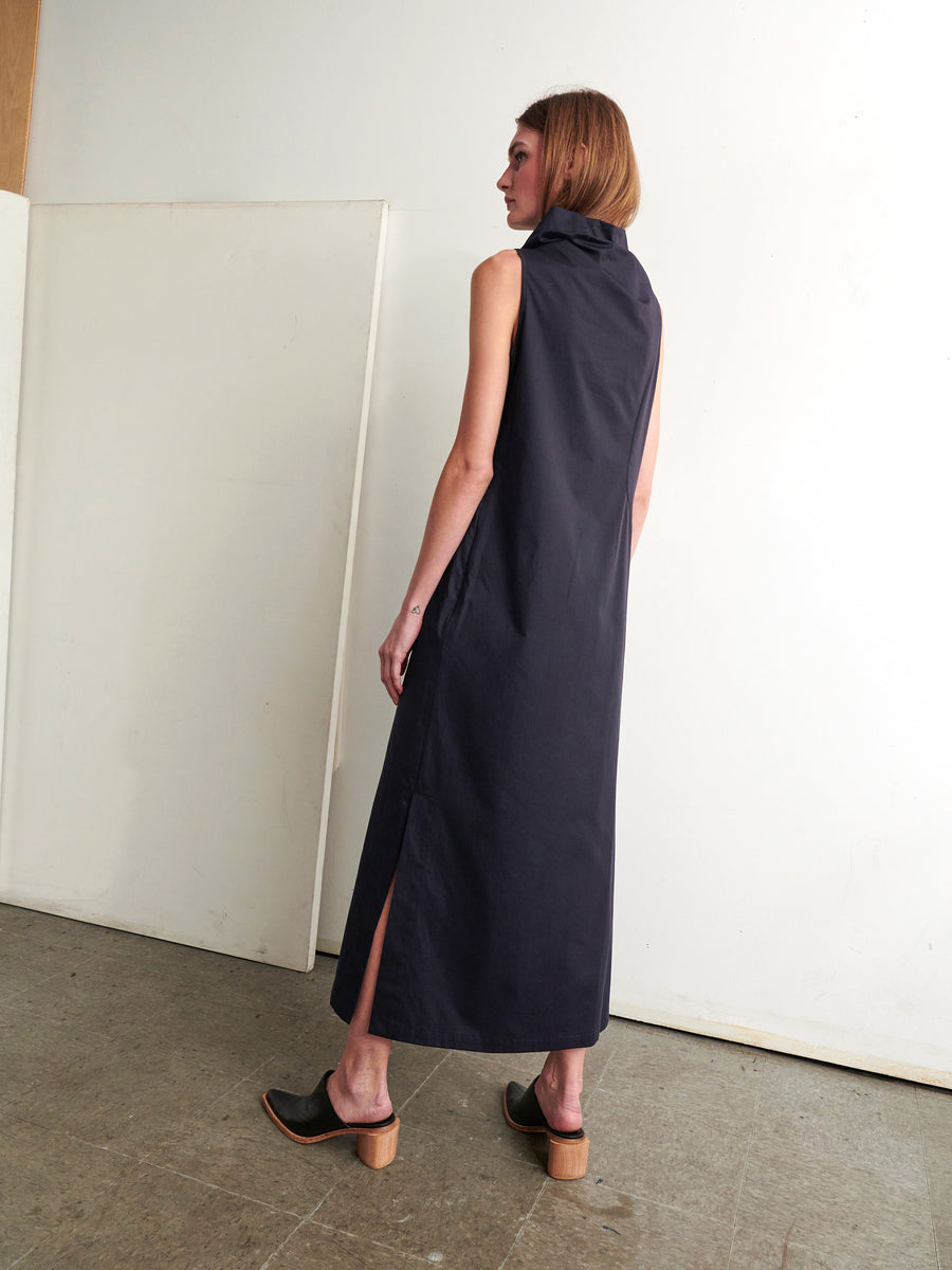 In a minimalist room, an individual with shoulder-length hair models the Long Fold Dress by Zero + Maria Cornejo. This sleeveless cotton dress features elegant asymmetrical detailing and side slits. The look is completed with black shoes featuring wooden heels, as they turn to the side to reveal the subtle sophistication of this New York-made piece.