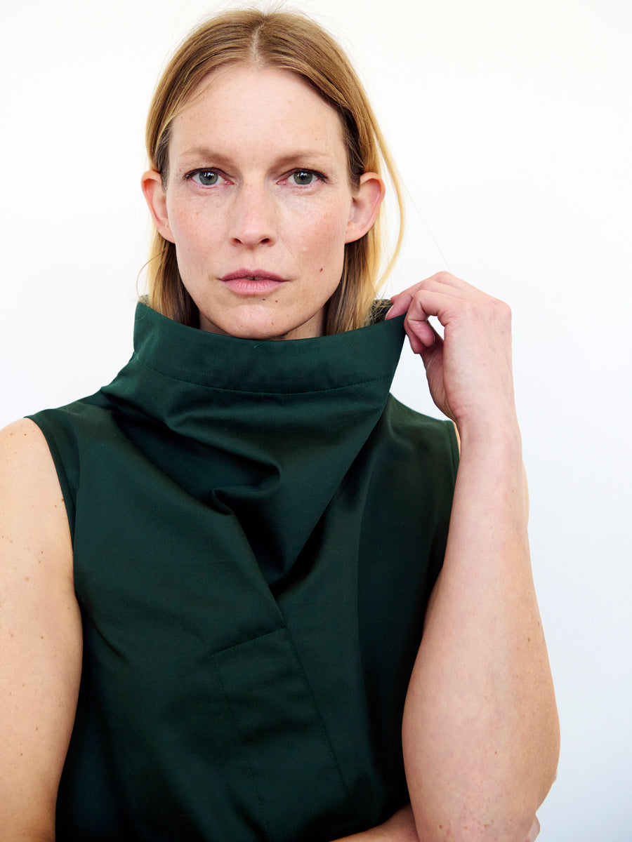 A person with long, light brown hair stares directly at the camera, holding up the collar of the Long Fold Dress from Zero + Maria Cornejo in dark green cotton broadcloth. The background is plain and light, highlighting the subject's serious expression and the asymmetrical details of this unique clothing design.