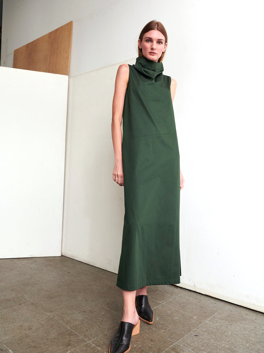 In a minimalist setting, a person is seen wearing the Long Fold Dress by Zero + Maria Cornejo—a sleeveless, floor-length dress crafted from dark green cotton broadcloth and featuring a high collar. They pose in profile with one arm at their side against a serene backdrop of neutral-toned flooring and simple, light-colored walls.