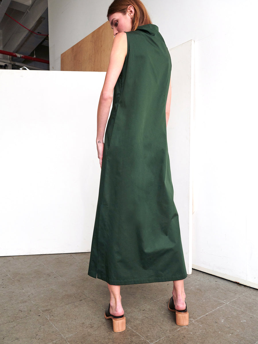 A person with long hair stands with their back to the camera, wearing the Long Fold Dress by Zero + Maria Cornejo, a sleeveless green dress made from cotton broadcloth featuring asymmetrical details. They pair it with brown platform sandals while standing in a room with white walls and a wooden panel.