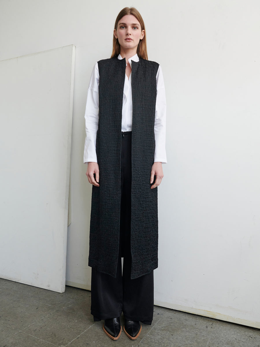 A person stands against a plain white wall, wearing a white collared shirt underneath the Luna Waistcoat by Zero + Maria Cornejo, expertly crafted from recycled polyester with a textured woven pattern. They are also dressed in black wide-leg pants and black pointy shoes. Their straight brown hair frames their face as they look directly at the camera.