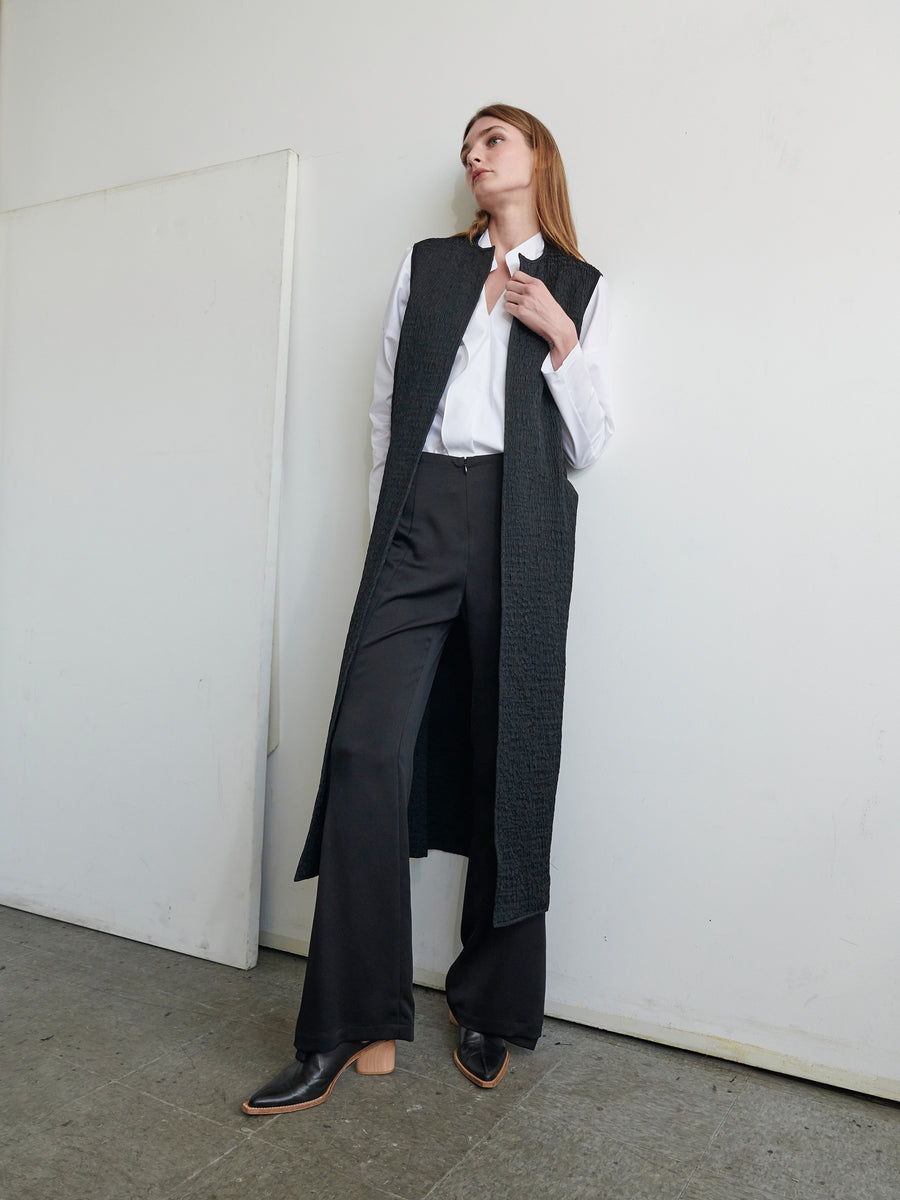 A person is leaning against a white wall, wearing the Luna Waistcoat by Zero + Maria Cornejo over a white shirt and black flared pants. They are looking upward with a relaxed expression. Their black heeled shoes with a tan sole complete the look in an indoor space.