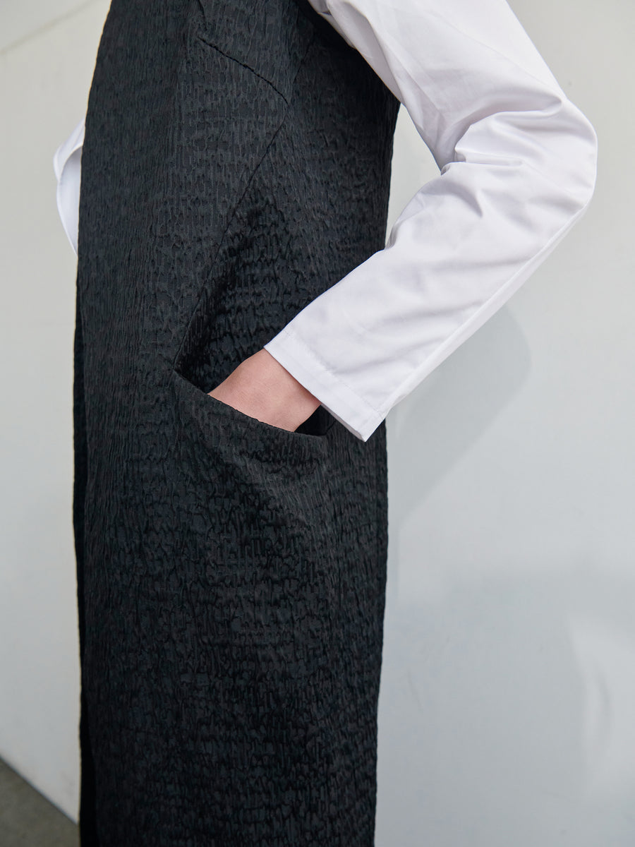 A person in a white long-sleeved shirt and the Luna Waistcoat, a textured black sleeveless dress made from recycled polyester by Zero + Maria Cornejo, is shown from the neck down. The person is placing their left hand into a pocket on the dress. The background is plain white.