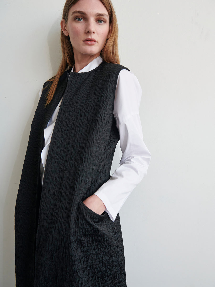 A woman with light skin and straight light-brown hair stands against a plain wall. She is wearing the Luna Waistcoat from Zero + Maria Cornejo over a white long-sleeve shirt made of organic cotton. Her left hand is in the waistcoat pocket, and she is gazing confidently at the camera.