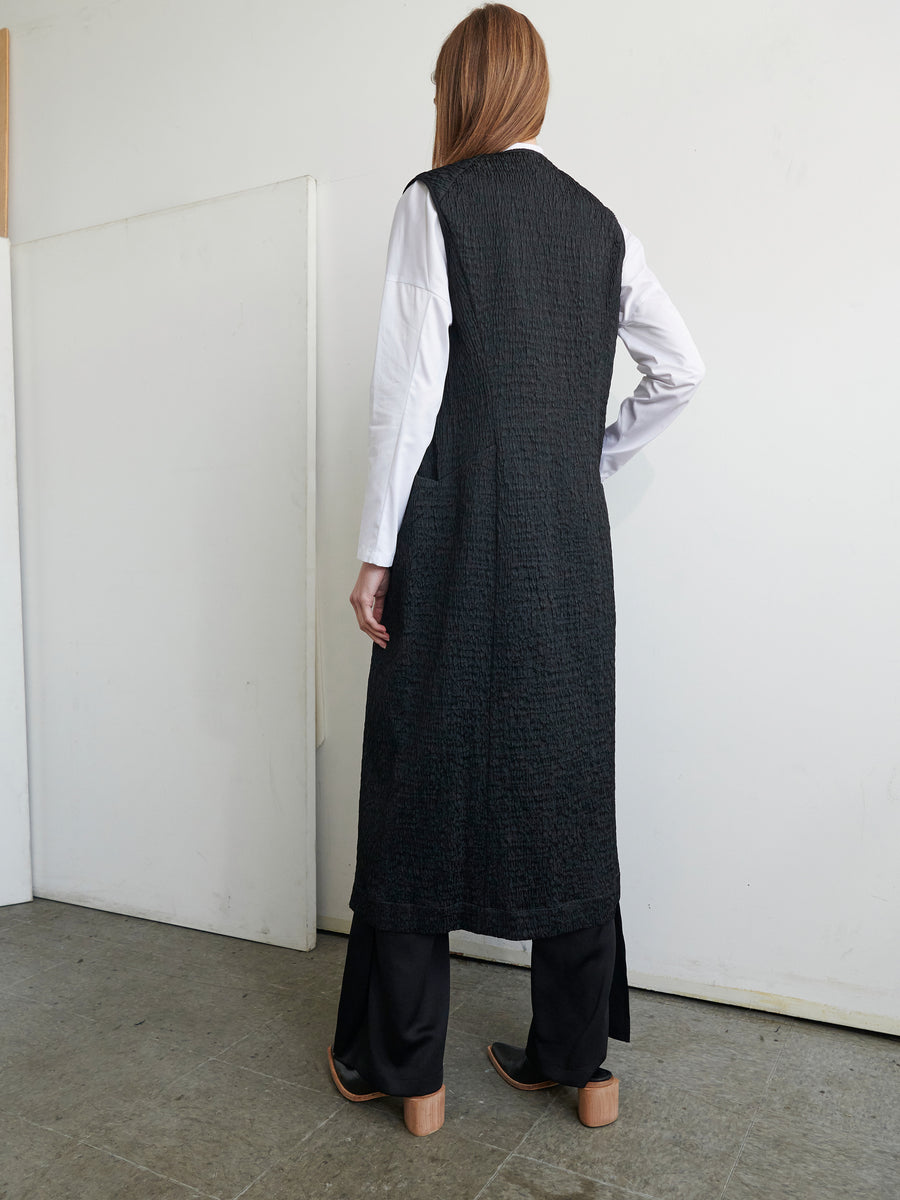 A person with long hair stands facing a white wall, wearing the Luna Waistcoat by Zero + Maria Cornejo, crafted from recycled polyester. They pair it with an organic cotton white shirt and black pants. Indoors, they stand on a gray concrete floor.