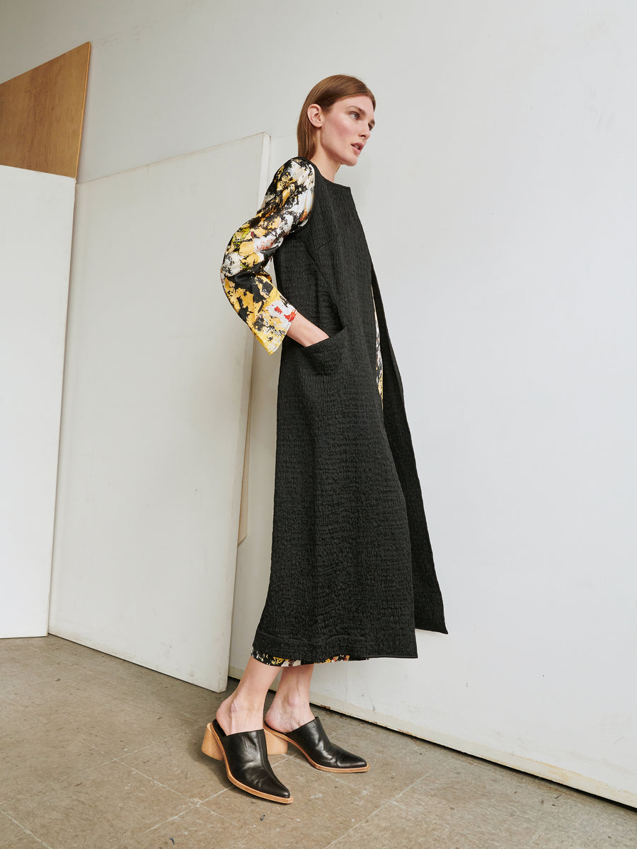 A person stands against a plain background, wearing the Luna Waistcoat, a sleeveless black textured long coat by Zero + Maria Cornejo, over a floral-patterned blouse and skirt. They have their hands in the coat pockets and are wearing black slip-on shoes with wooden heels. The person gazes to their left, showcasing an ensemble that includes pieces made from recycled polyester.