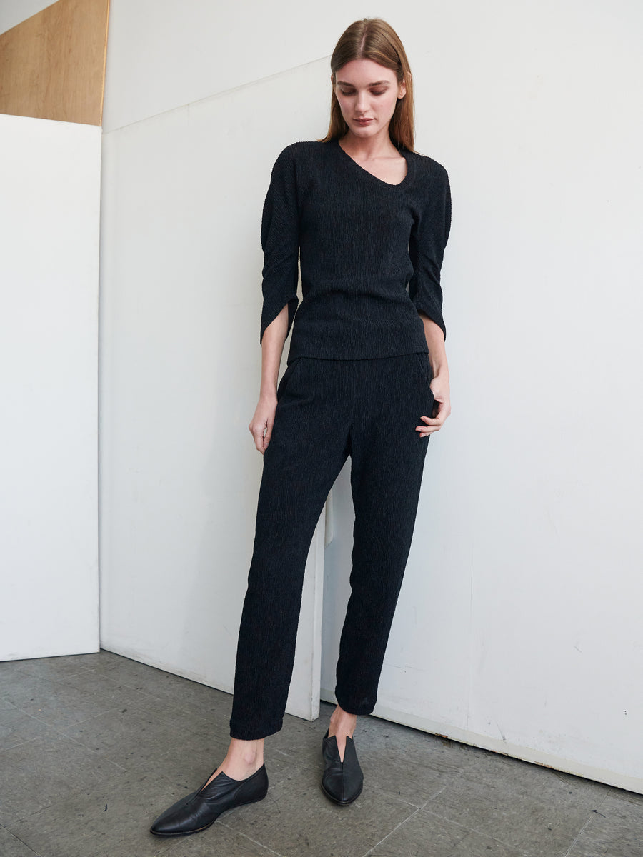 A woman stands against a white wall, dressed in a black long-sleeve top and Zero + Maria Cornejo's Gabi Trousers made from eco fabric. She has her hands in her pockets and wears black slip-on shoes. She looks down slightly, with one foot slightly forward on the grey, textured surface floor.