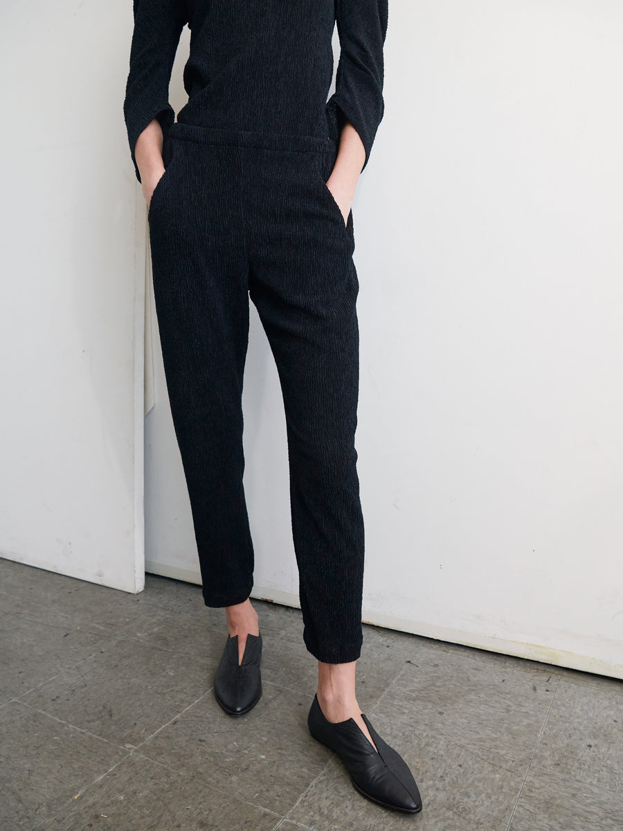 A person stands against a white wall, wearing a black long-sleeve top and the Gabi Trouser by Zero + Maria Cornejo. Their hands are in their pockets, paired with black slip-on shoes. The floor is gray with some texture. The person's face is not visible.