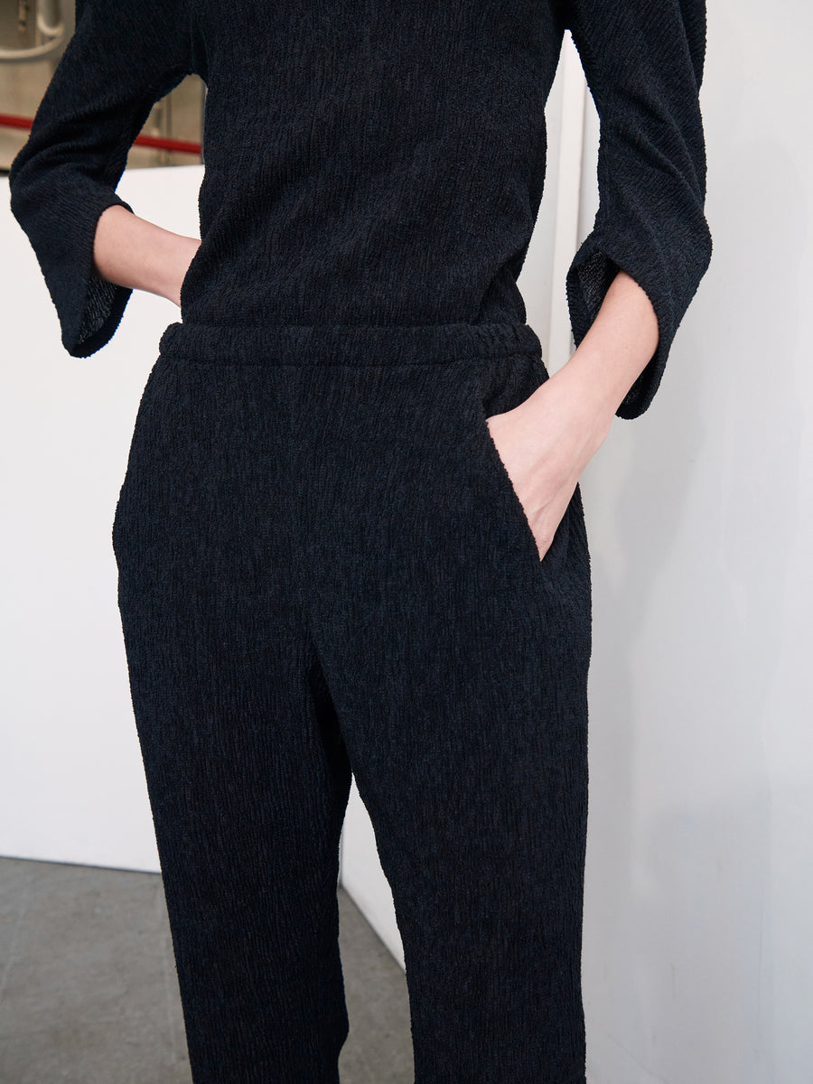 A person wearing a black, long-sleeved top and the Gabi Trouser, designed by Zero + Maria Cornejo and made from recycled polyester, is standing with one hand tucked into a pants pocket. The background reveals a minimalist indoor setting with white walls and a hint of a red line.