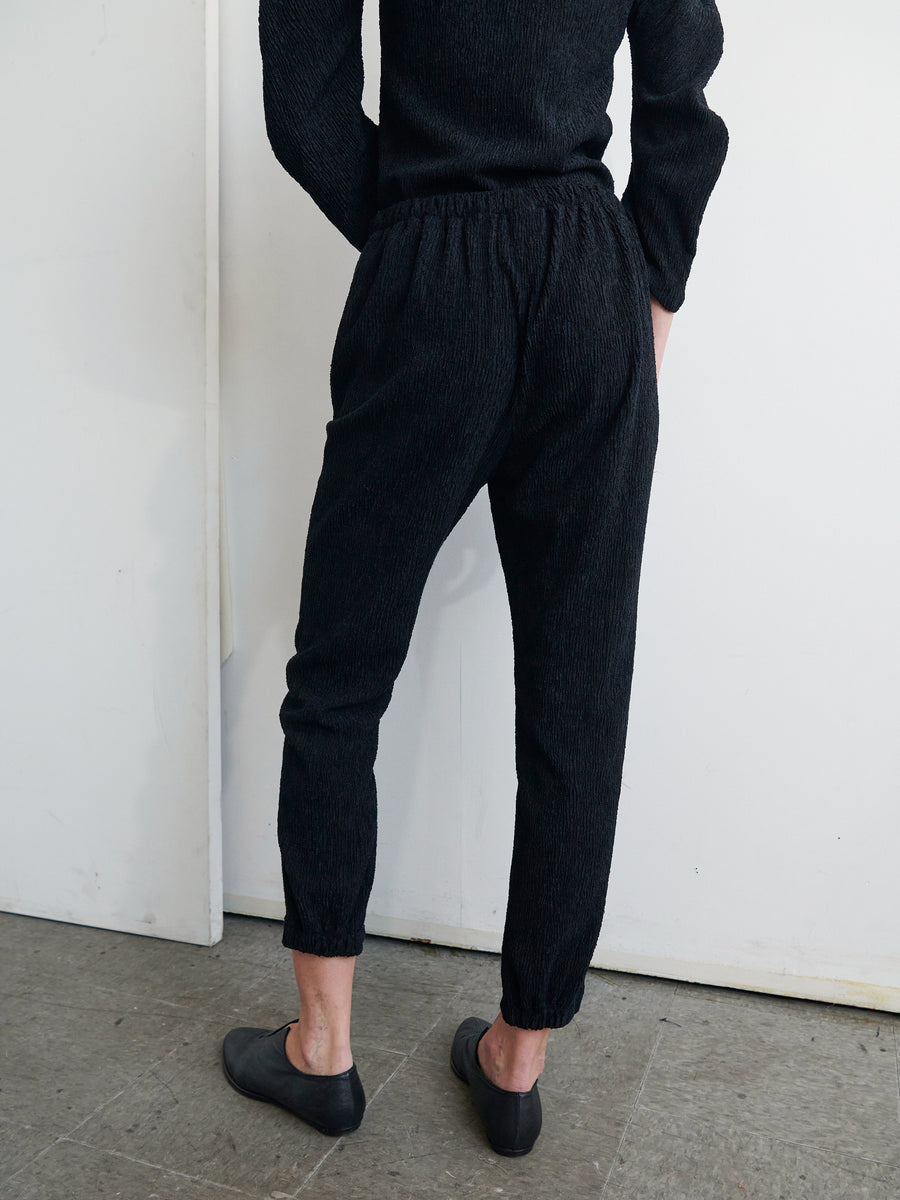 A person stands facing away, wearing a black long-sleeve top, the Gabi Trouser in black stretch boucle eco fabric by Zero + Maria Cornejo, and black shoes. They are positioned against a plain white wall on a gray textured floor. The outfit appears casual and comfortable.