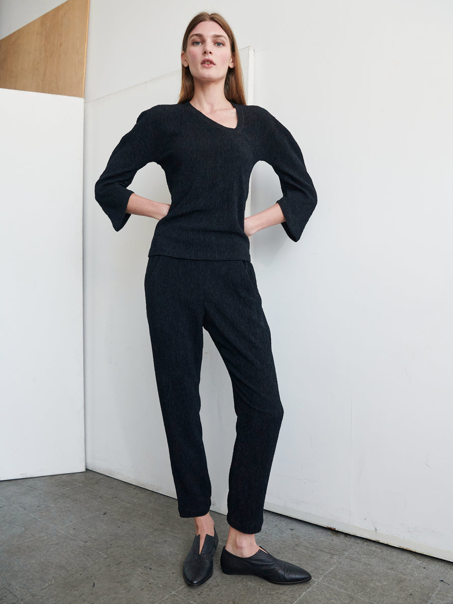 A person with long brown hair stands against a white wall on a gray floor. They are wearing the Long-Sleeved Off Kilter Top from Zero + Maria Cornejo in charcoal gray, paired with matching eco boucle stretch fabric pants and black slip-on shoes. Their hands rest on their hips, and they are gazing slightly downward.