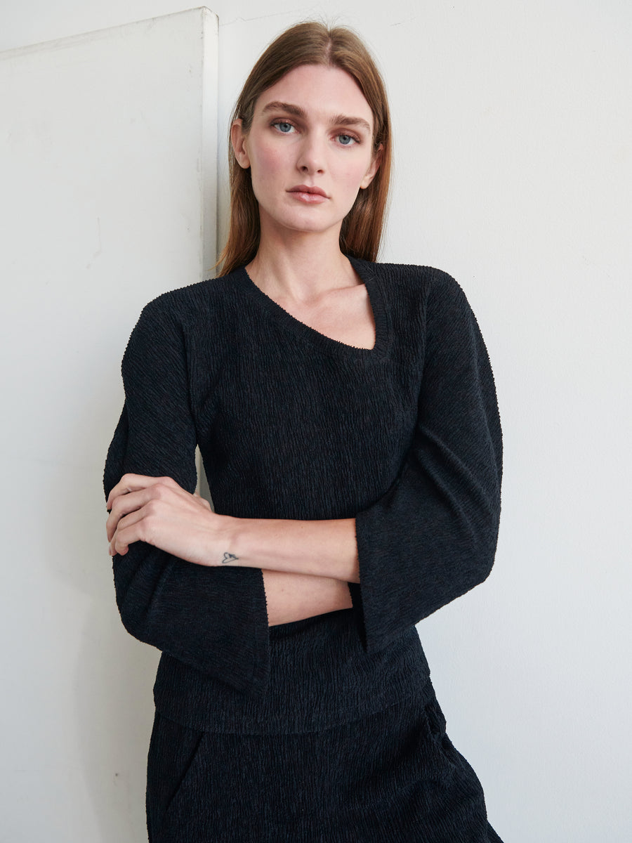 A woman with long brown hair is standing with her arms crossed, wearing a black Long-Sleeved Off Kilter Top by Zero + Maria Cornejo. She is positioned against a light-colored wall and is looking straight into the camera with a neutral expression.