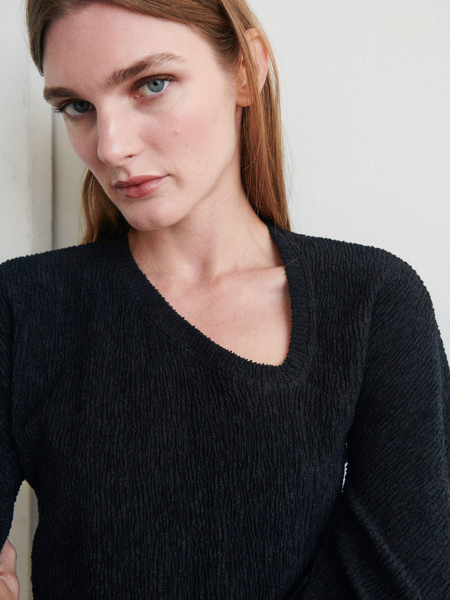 A person with long, light brown hair looks directly at the camera with a neutral expression. They are wearing a Long-Sleeved Off Kilter Top from Zero + Maria Cornejo, made from eco boucle stretch fabric. The background is a plain, light-colored wall.