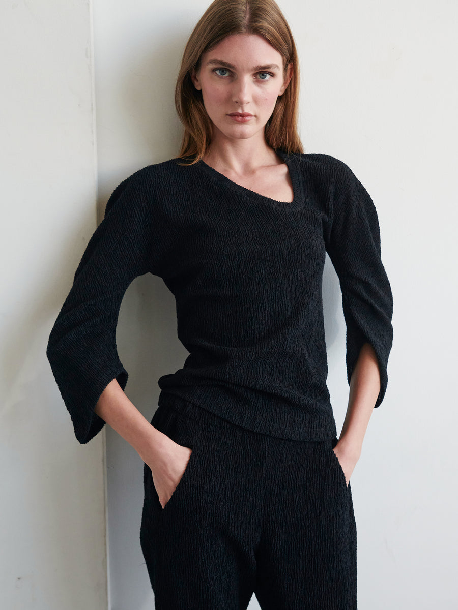 A woman with long hair stands against a light wall, wearing Zero + Maria Cornejo’s Long-Sleeved Off Kilter Top and matching black pants. She has her hands in her pockets and gazes directly at the camera with a neutral expression. The recycled polyester top features a distinctive neckline and three-quarter length sleeves.