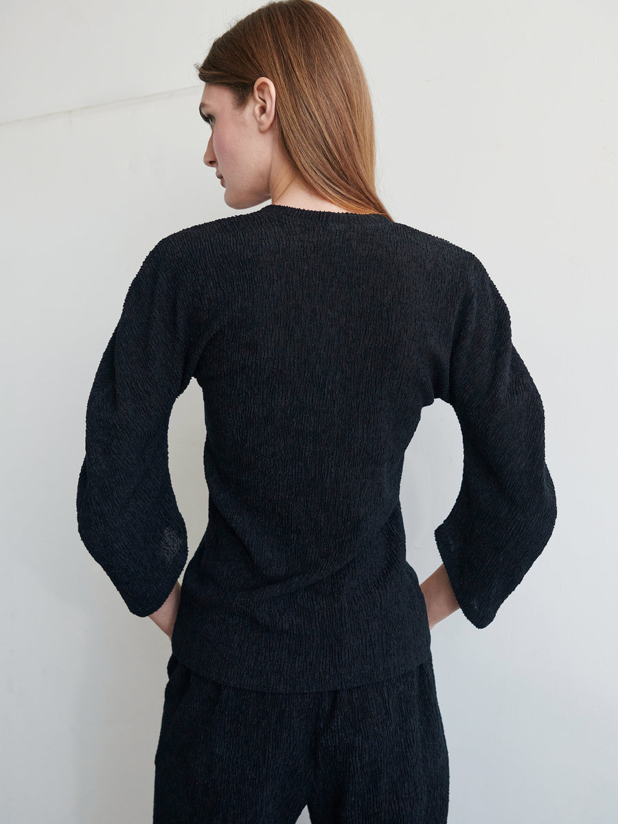 A woman with long, reddish hair stands facing away from the camera, showcasing the back of her outfit. She is wearing a Black Long-Sleeved Off Kilter Top and matching pants made from recycled polyester by Zero + Maria Cornejo. The background is off-white and minimalistic.