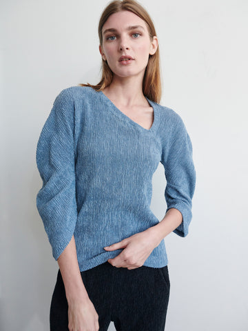 A person with long light brown hair stands against a plain background, wearing the Long-Sleeved Off Kilter Top in blue from Zero + Maria Cornejo made from eco boucle stretch fabric and black pants. The individual gazes at the camera with a neutral expression, one hand resting on their side.