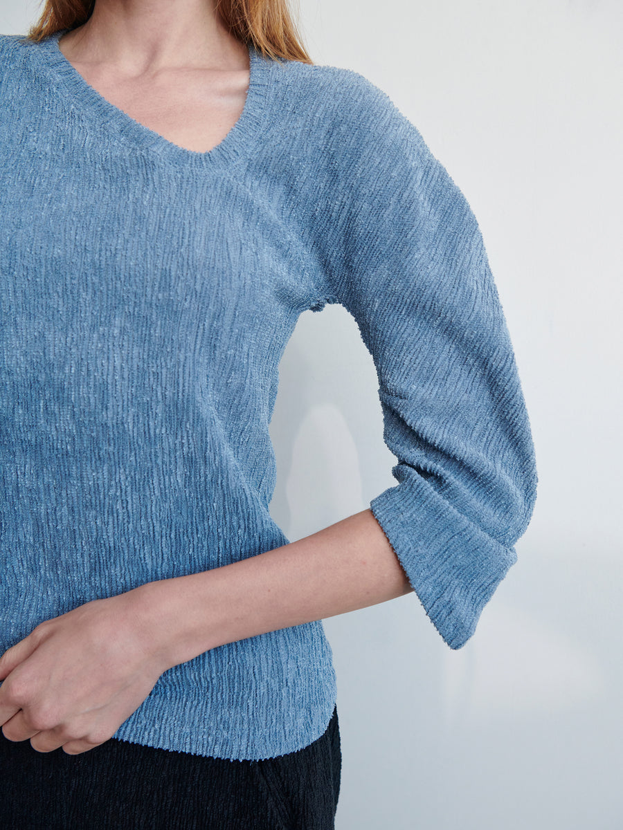 A person with long hair is wearing a light blue, textured top called the Long-Sleeved Off Kilter Top by Zero + Maria Cornejo. The top has a V-neck and three-quarter length sleeves. The person is seen from the chest to the hips, one arm bent at the elbow. The background is plain and light-colored.