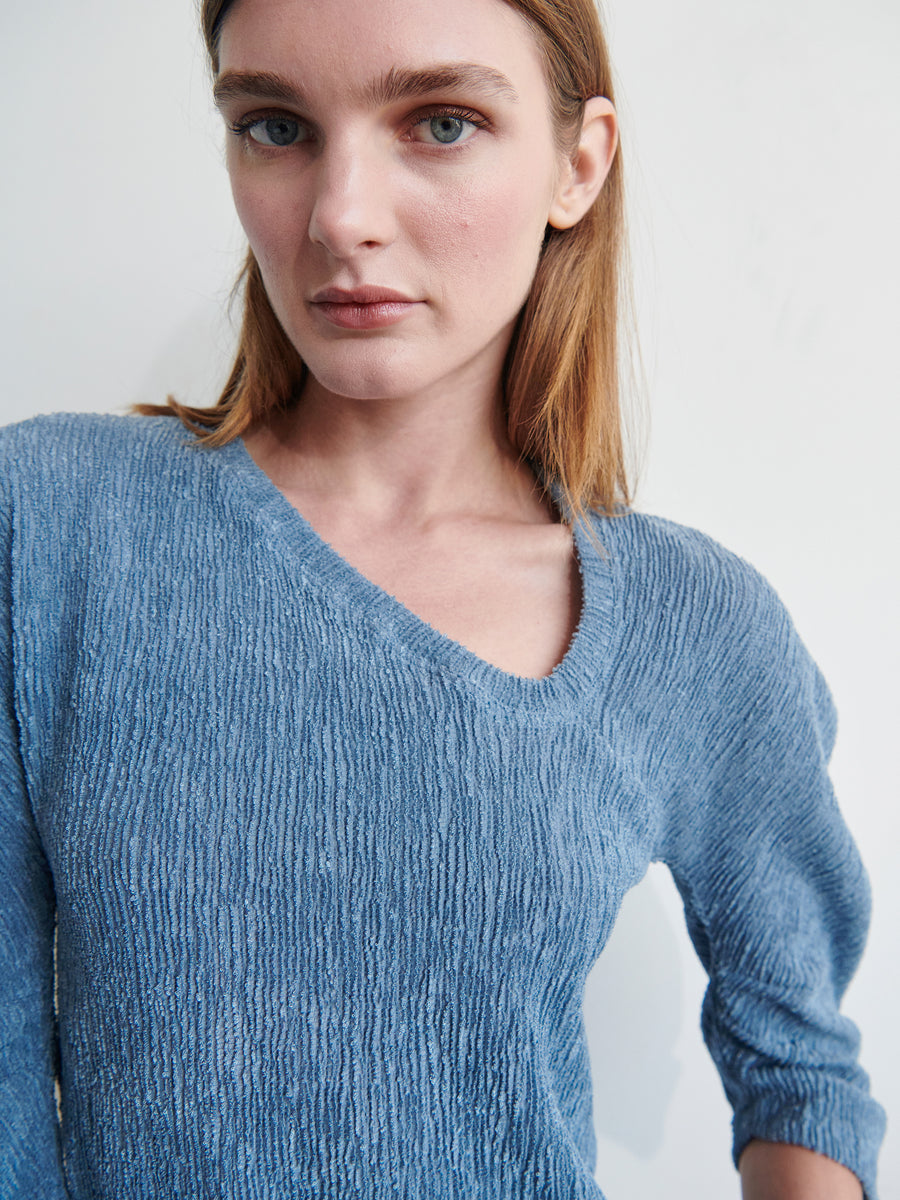 A woman with straight, light brown hair and blue eyes is looking directly at the camera. She is wearing a blue Long-Sleeved Off Kilter Top made from recycled polyester by Zero + Maria Cornejo and standing against a plain white background. The lighting is soft, highlighting her facial features and the top's texture.