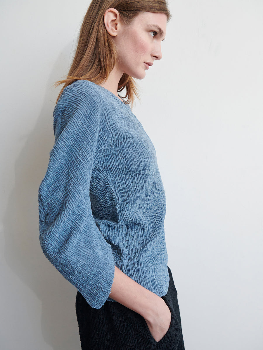 A person with shoulder-length hair is posing sideways against a plain white background. They are dressed in a textured blue Long-Sleeved Off Kilter Top by Zero + Maria Cornejo and dark trousers, with one hand in their pocket. The lighting highlights the texture of the clothing.