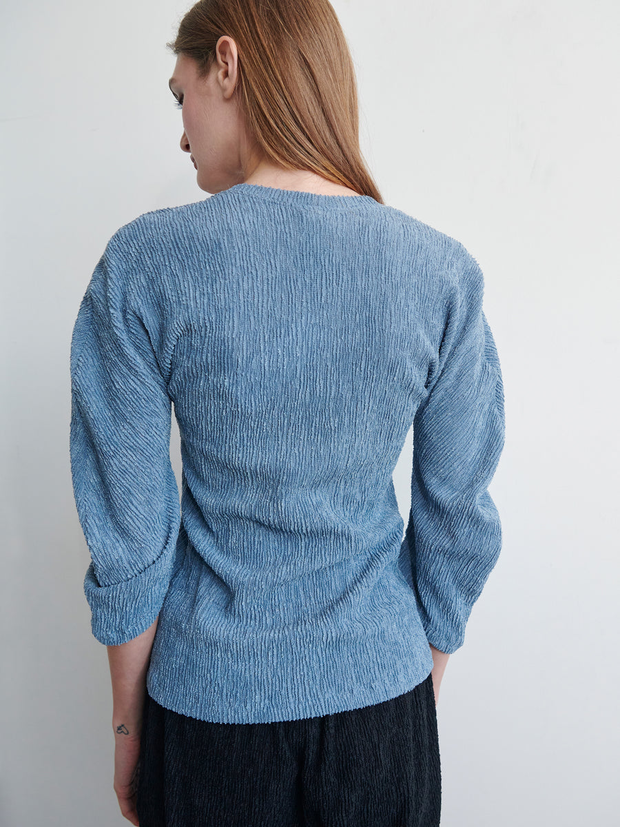 A person with long brown hair is seen from the back, wearing a Long-Sleeved Off Kilter Top by Zero + Maria Cornejo made of eco boucle stretch fabric, paired with dark pants. The soft lighting highlights the texture of the clothing. The background is plain, featuring a light, neutral-colored wall.