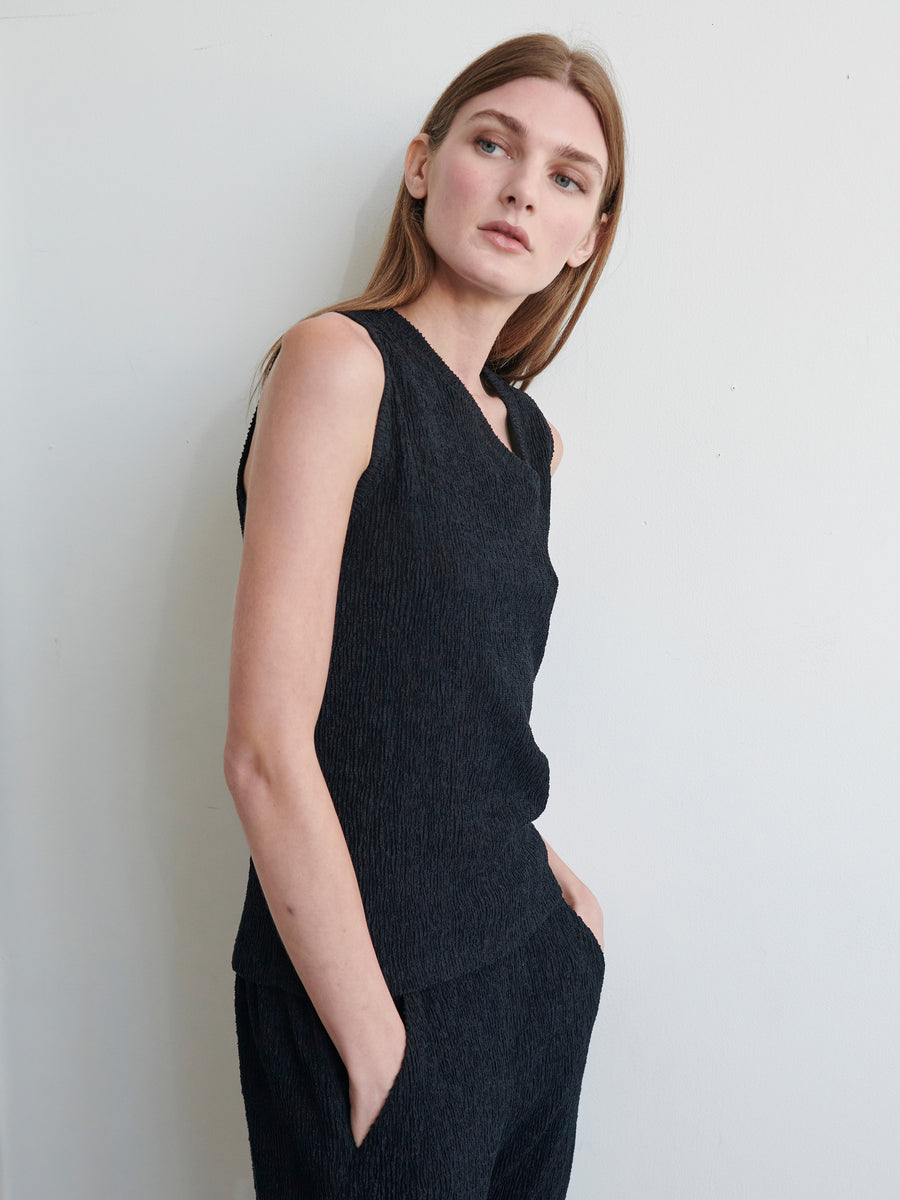 A person with long, straight hair stands against a plain white background, wearing the Off Kilter Tank from Zero + Maria Cornejo with its asymmetrical neckline made from eco boucle fabric, paired with matching black pants. They have their left hand in a pocket and look slightly to their right, exuding a composed expression.