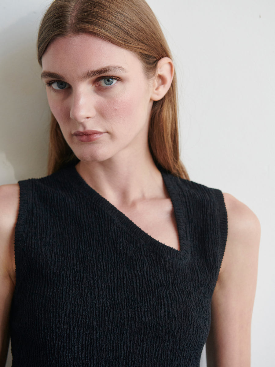 A woman with long, straight hair and light eyes is standing against a plain background, gazing slightly to the side. She is wearing the Off Kilter Tank from Zero + Maria Cornejo, a sleeveless top made of textured stretch eco boucle fabric featuring a V-neckline. Her expression is neutral.