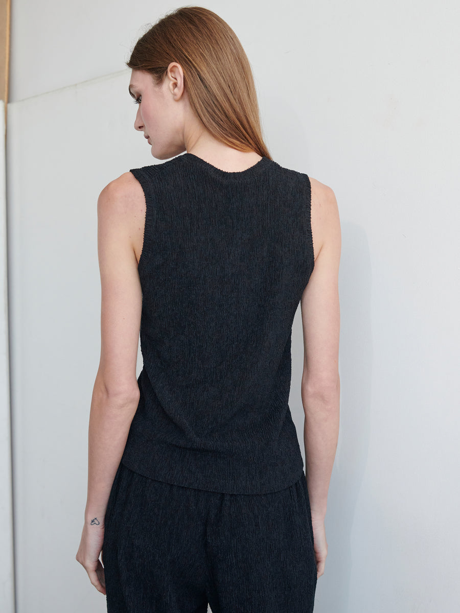 A person with long brown hair is standing with their back to the camera. They are wearing the Off Kilter Tank by Zero + Maria Cornejo, a black, textured stretch sleeveless top, paired with matching black pants made from recycled polyester. The background is plain and light-colored as their head is slightly turned to the side.