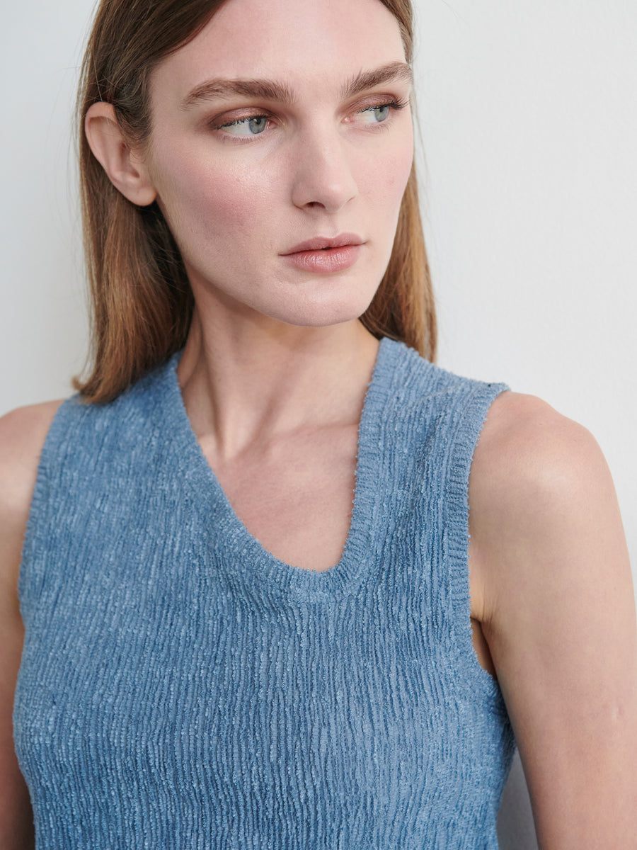 A light-skinned individual with long brown hair is shown from the shoulders up, wearing the Off Kilter Tank by Zero + Maria Cornejo. The sleeveless top, featuring a blue textured stretch design made of eco boucle stretch fabric, complements their neutral expression as they look to the left against a plain, light-colored background.