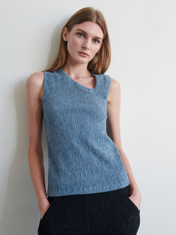 A person with long, straight hair, wearing the Off Kilter Tank by Zero + Maria Cornejo in blue, paired with black pants, stands against a white wall. They have a relaxed posture with their hands in their pockets, looking directly into the camera.