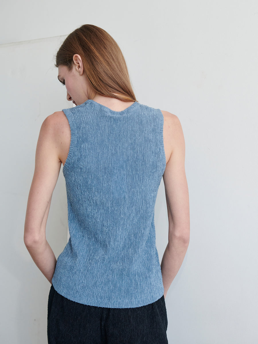 A person with long brown hair is standing with their back to the camera, wearing the Off Kilter Tank by Zero + Maria Cornejo in blue and dark trousers. The background is plain and light-colored, highlighting the eco boucle stretch fabric of the outfit.