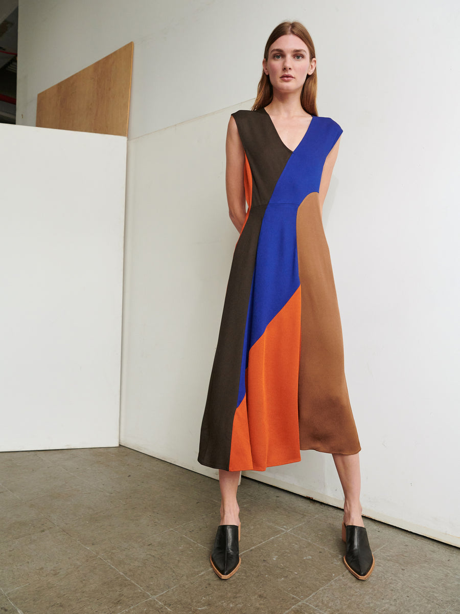 A woman stands indoors against a minimalist background wearing the Eve Mosa Dress from Zero + Maria Cornejo, a sleeveless, color-block mid-length dress in shades of black, blue, orange, and brown. Made from sustainable viscose, she has long brown hair and is wearing black slip-on shoes. Her hands are clasped behind her back as she looks ahead.