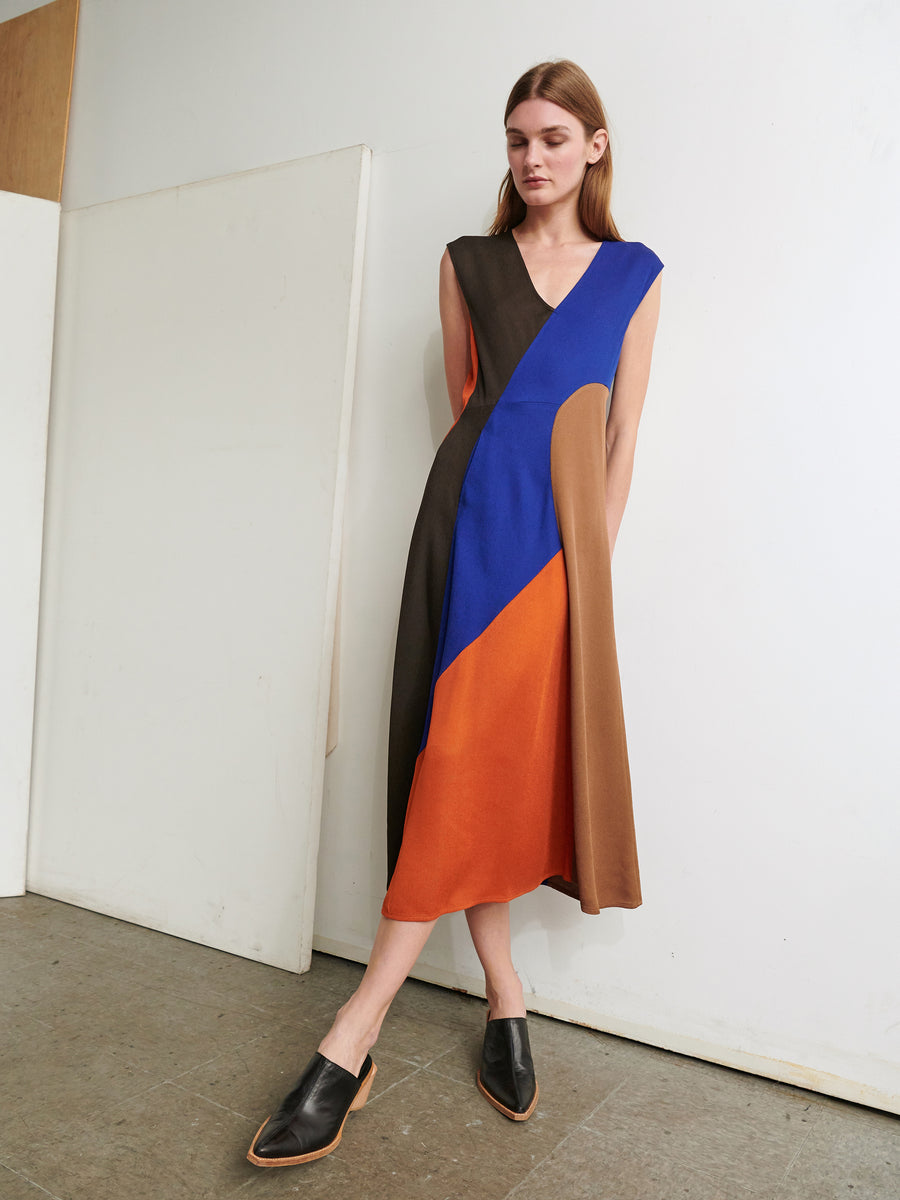 A woman with straight, light brown hair stands against a white wall, wearing the Eve Mosa Dress from Zero + Maria Cornejo, adorned in colorblocks of black, blue, orange, and tan. Embracing sustainable fashion, she pairs the recycled drape dress with black slip-on shoes and looks down with her hands behind her back.