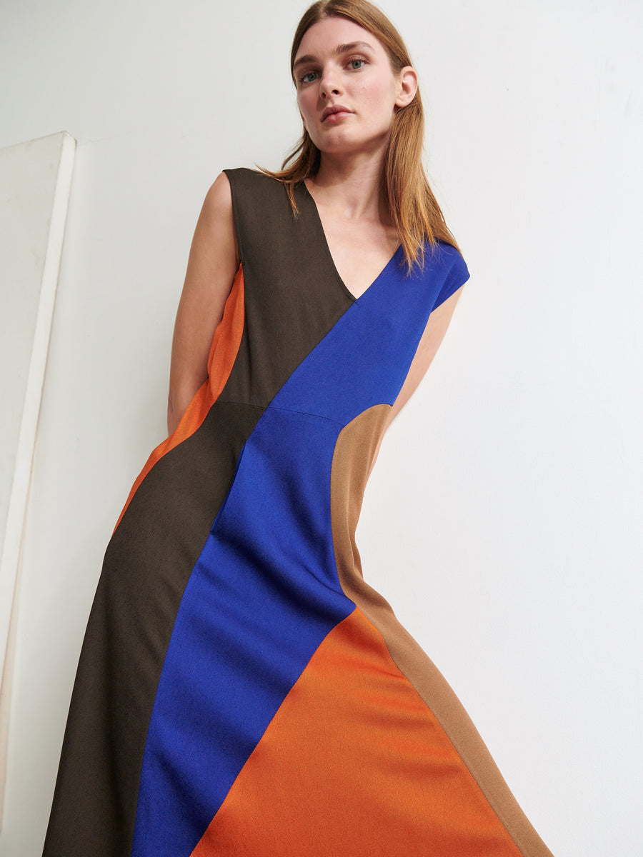 A person with long, straight hair is wearing the Eve Mosa Dress by Zero + Maria Cornejo. The sleeveless dress features bold geometric color blocks in brown, blue, black, and orange. They are standing against a plain white wall, looking slightly to the side, embodying the essence of sustainable fashion.