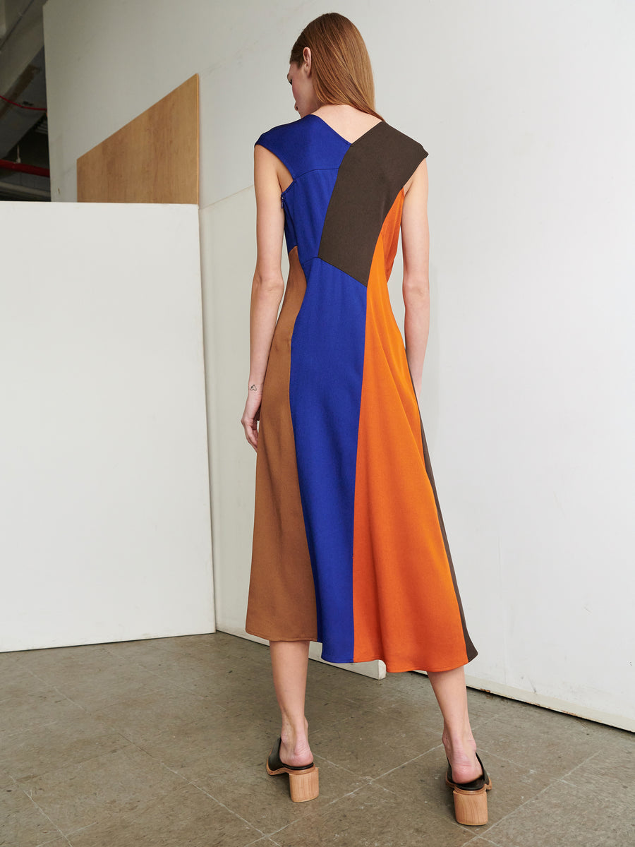 A woman with long hair stands indoors, wearing the Eve Mosa Dress by Zero + Maria Cornejo. The sleeveless, asymmetrical color-blocked mid-length dress features shades of blue, brown, and orange. Facing away from the camera, she is set against a plain white wall and wooden panel. Her open-toed shoes complete the look.