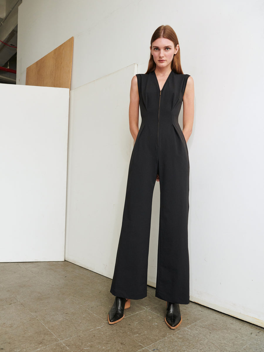 A woman stands against a white wall wearing the Chic Jumpsuit by Zero + Maria Cornejo, which features a black sleeveless design with a cut-out back and a front zipper. She has straight brown hair and is looking directly at the camera, completing her look with black heeled shoes.