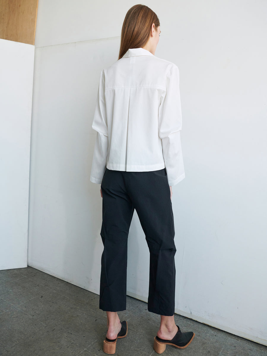 A person with long hair stands indoors, their back to the camera. They are wearing a white long-sleeved shirt and Zero + Maria Cornejo's Biker Pant, which features stretch woven fabric and front zip pockets, paired with black slip-on clogs with brown soles. The floor is concrete, and the walls are white.