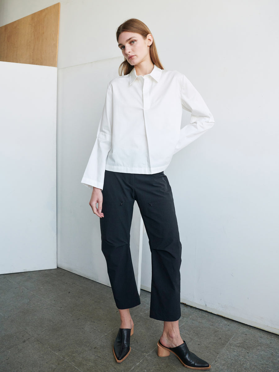 A person is standing indoors near a white and wooden wall, wearing a white long-sleeve shirt with bell sleeves, black Zero + Maria Cornejo Biker Pants, and black heeled mules. The individual has long hair and is posing with one hand on their hip and one leg slightly bent.