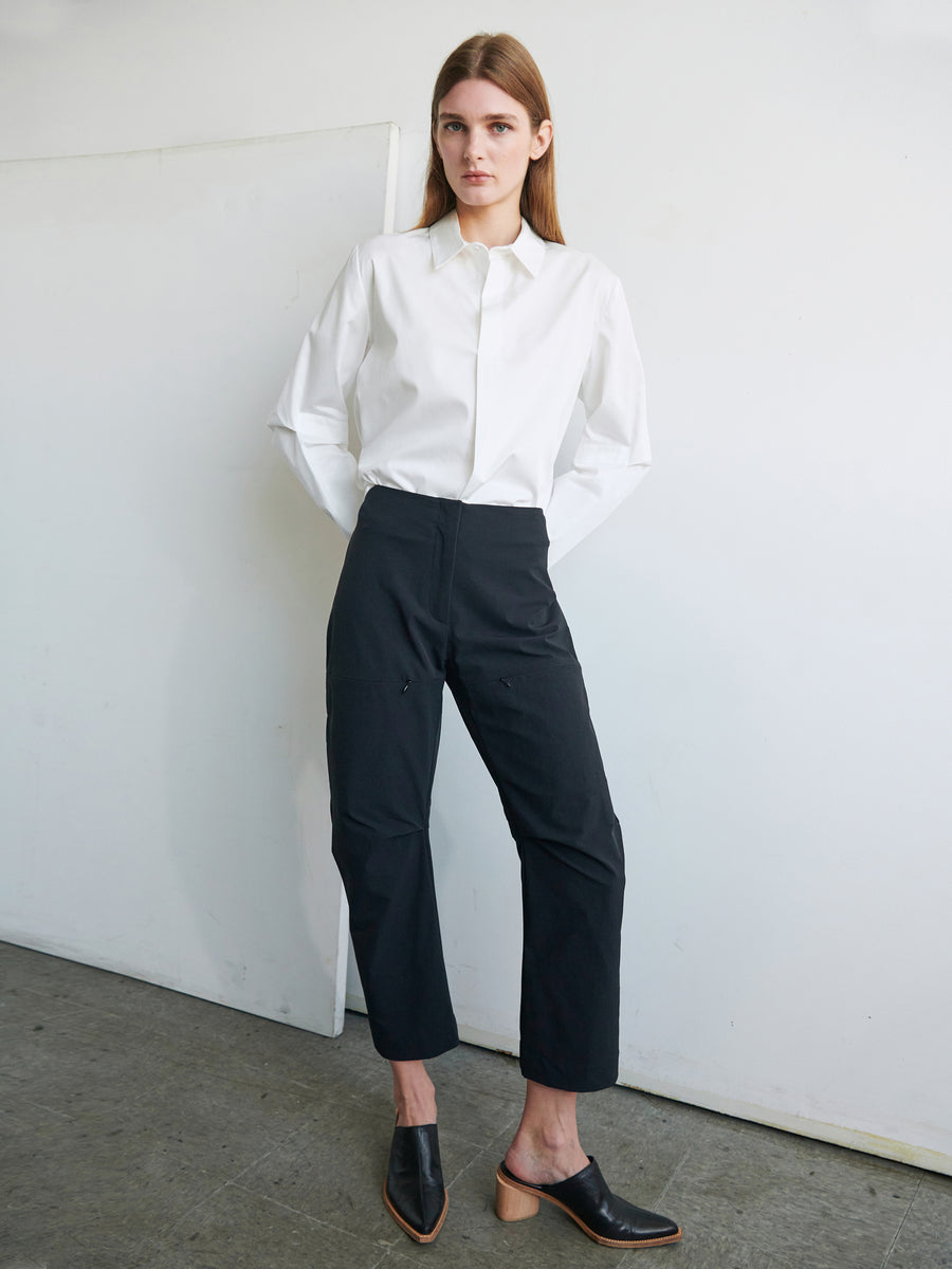 A person with long, straight hair is standing against a white wall. They are wearing a white collared shirt with long sleeves and black Biker Pant by Zero + Maria Cornejo. Their hands are behind their back, and they have on black shoes featuring a small wooden heel.