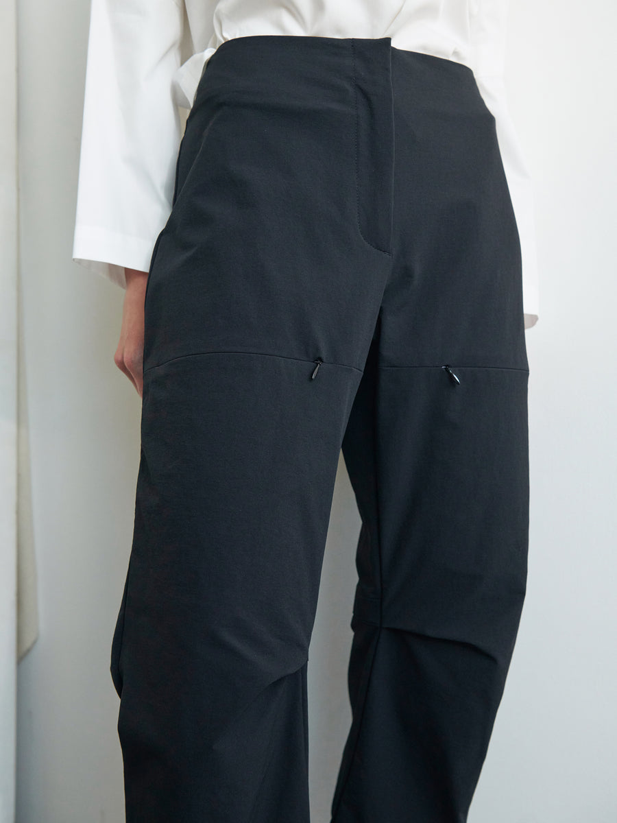 Close-up of a person wearing black Zero + Maria Cornejo Biker Pants with two small black drawstrings at the thigh area. The comfy structured design pairs perfectly with a white long-sleeve top. This premium New York-made product is set against a plain and neutral background.