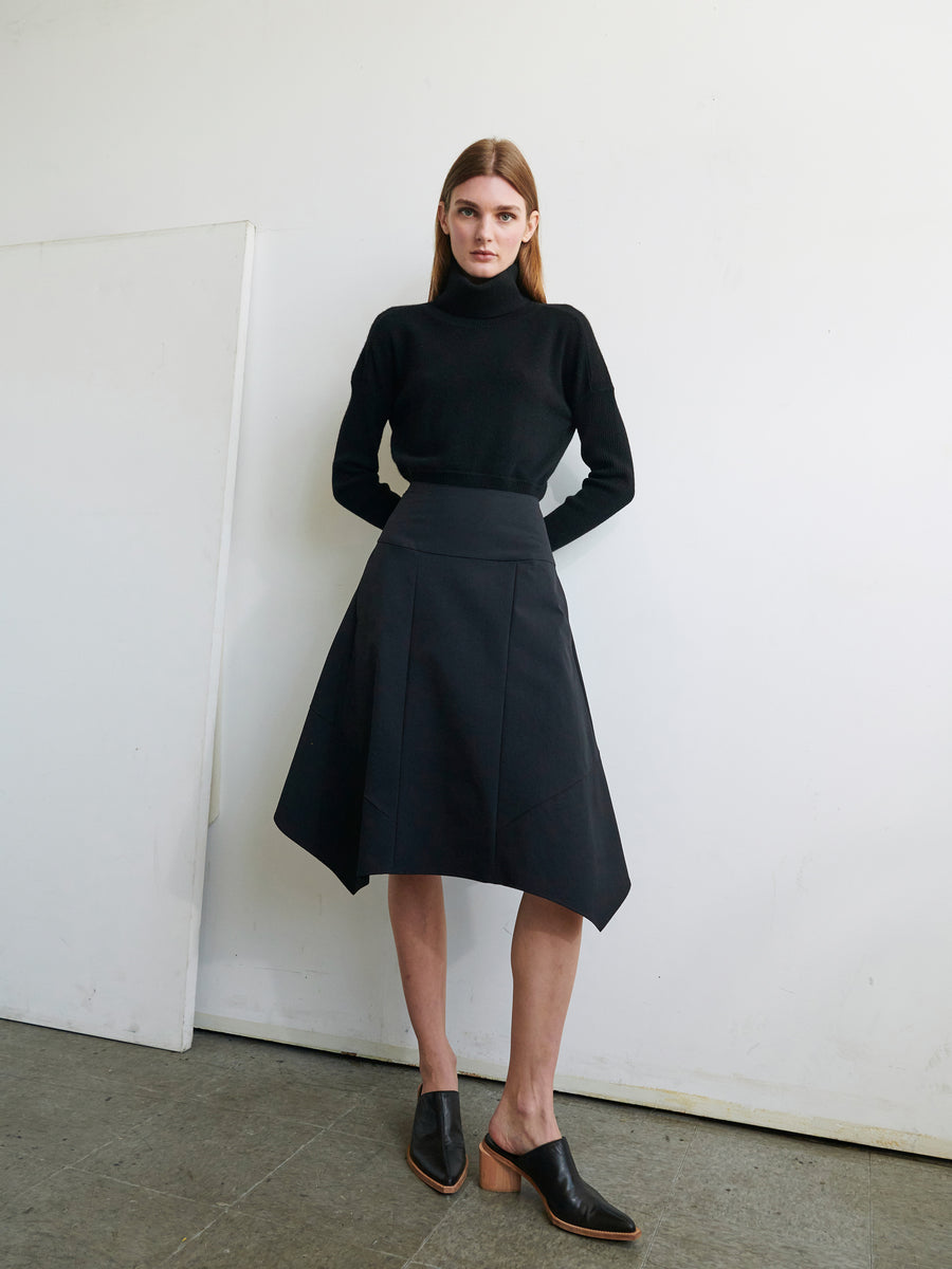 A woman with long hair stands against a white wall, wearing a black turtleneck sweater and the Madi Skirt by Zero + Maria Cornejo, featuring an asymmetrical hemline made of technical fabric. She has her hands clasped behind her back and is wearing black mules with wooden heels. The floor is concrete, and there's a white panel leaning against the wall.