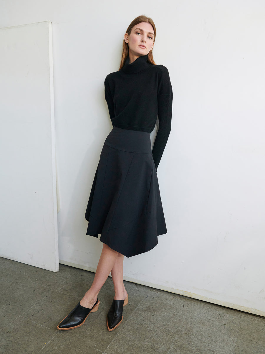 A person with long hair stands against a white wall. They are wearing a black turtleneck sweater, the Zero + Maria Cornejo Madi Skirt with an asymmetrical hemline, and black pointed-toe shoes. The person is looking slightly off-camera with a neutral expression.