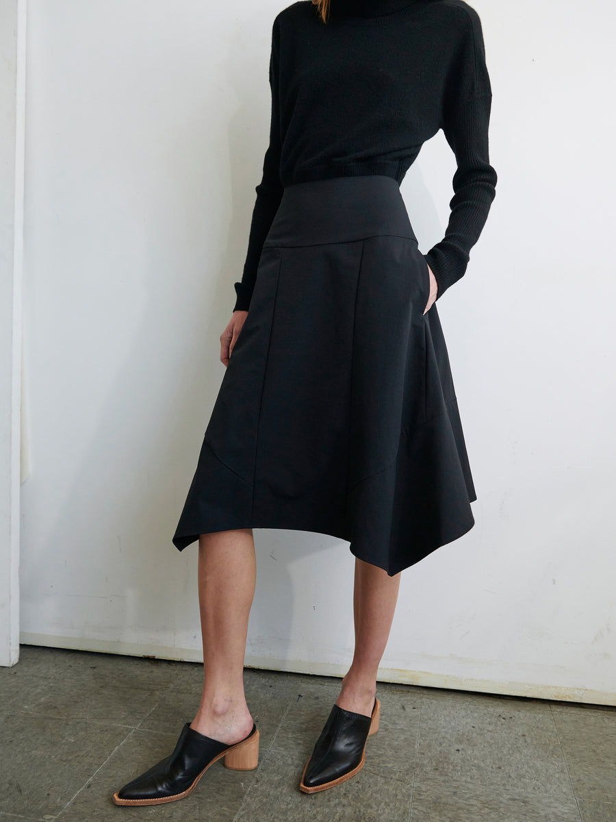 A person is wearing a black turtleneck sweater and the Madi Skirt from Zero + Maria Cornejo, an asymmetrical stretch-woven skirt with pockets. They are also wearing black leather mules with a wooden block heel. The background features a plain white wall and a gray tiled floor. The person's head is out of frame.