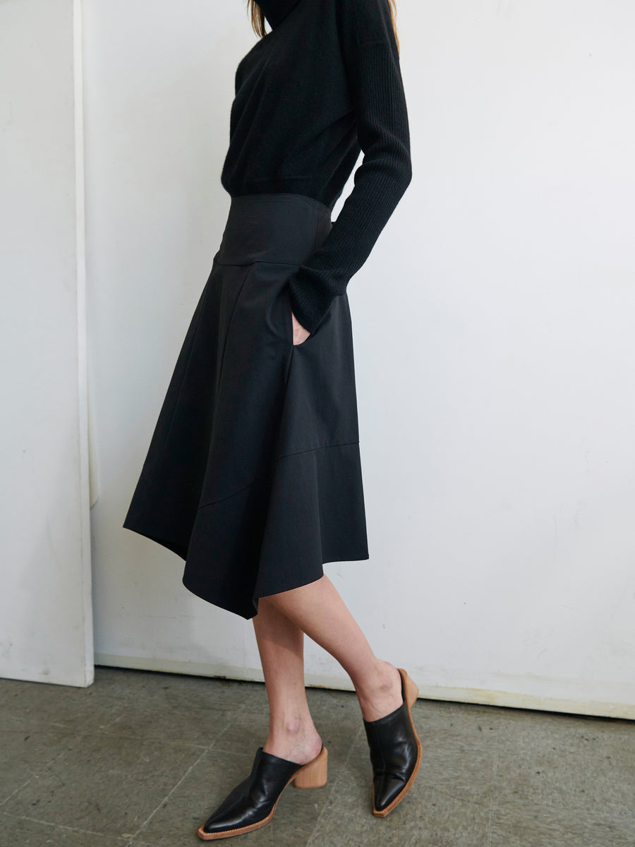 A woman is standing against a white wall. She is wearing a black long-sleeve top, the Madi Skirt by Zero + Maria Cornejo, which is a black stretch-woven skirt with pockets, and black mules with wooden block heels. The floor is gray, and her hands are in her skirt pockets.