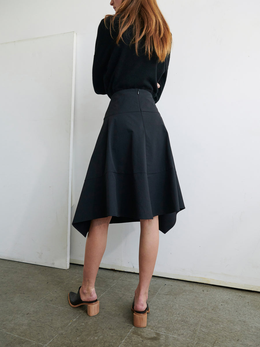 A person with long hair, seen from the back, is wearing a black long-sleeve top and the Madi Skirt by Zero + Maria Cornejo, featuring an asymmetrical hemline made from technical fabric. They are standing in a minimalistic indoor space with light-colored walls and a concrete floor, and are wearing black open-back sandals with wooden heels.